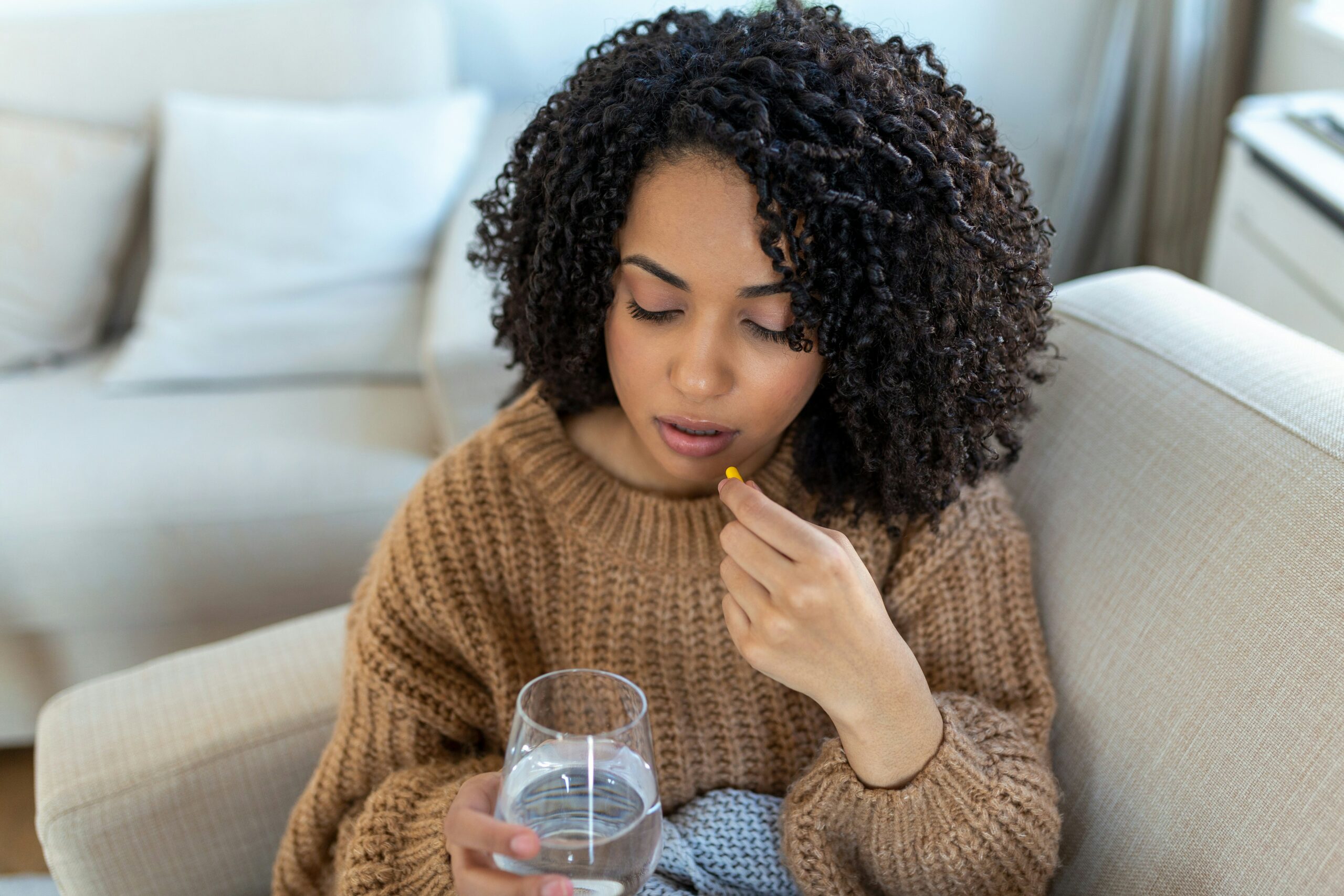 a young black woman sitting on her couch taking a pill with water